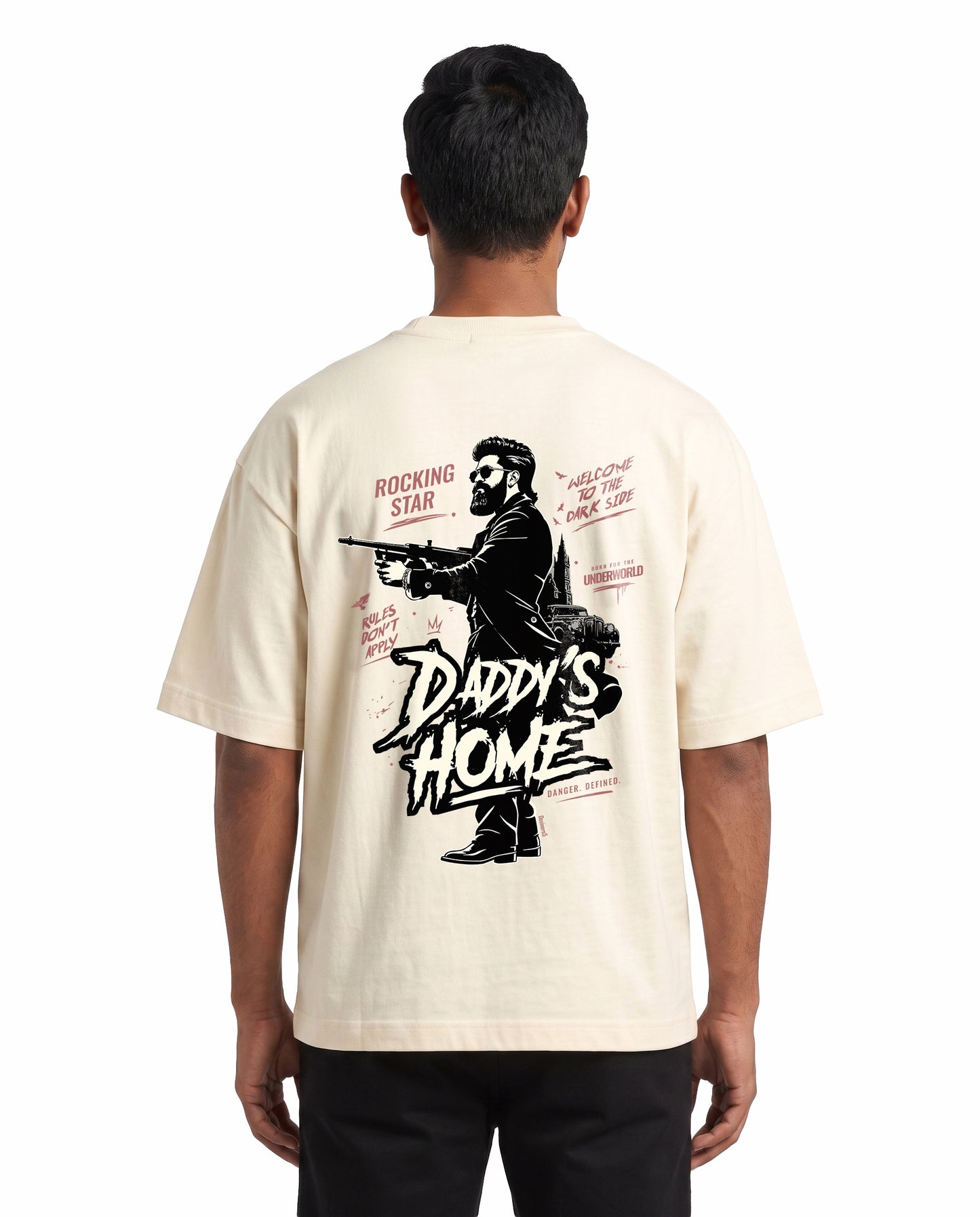 Back view of male model wearing DADDY'S HOME unisex oversized graphic t-shirt in off-white in a studio setting.