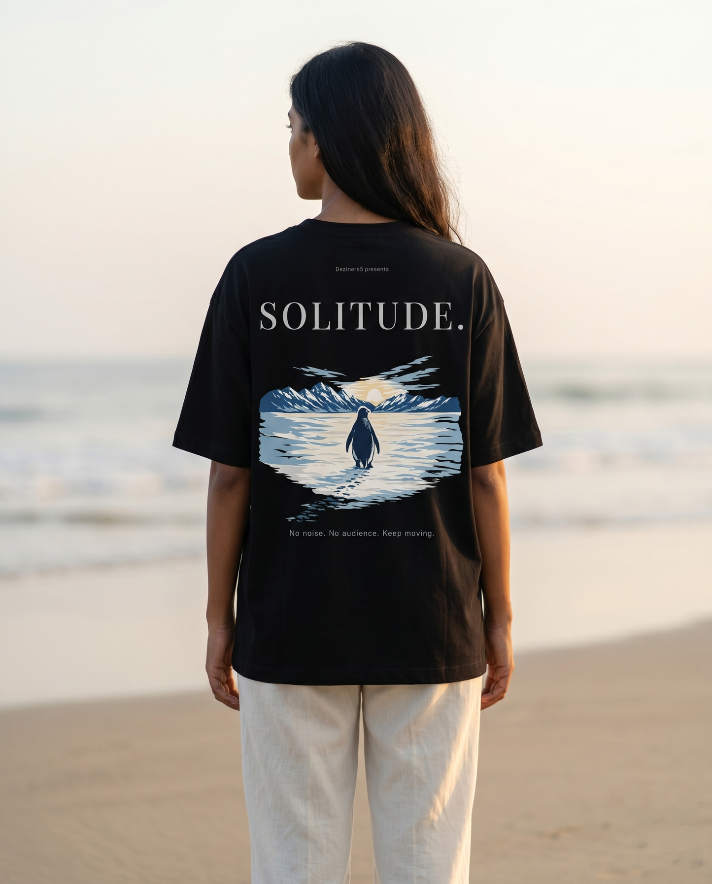 South Indian woman wearing the SOLITUDE - unisex oversized graphic t-shirt in black on a beach at sunrise featuring inspirational penguin artwork.