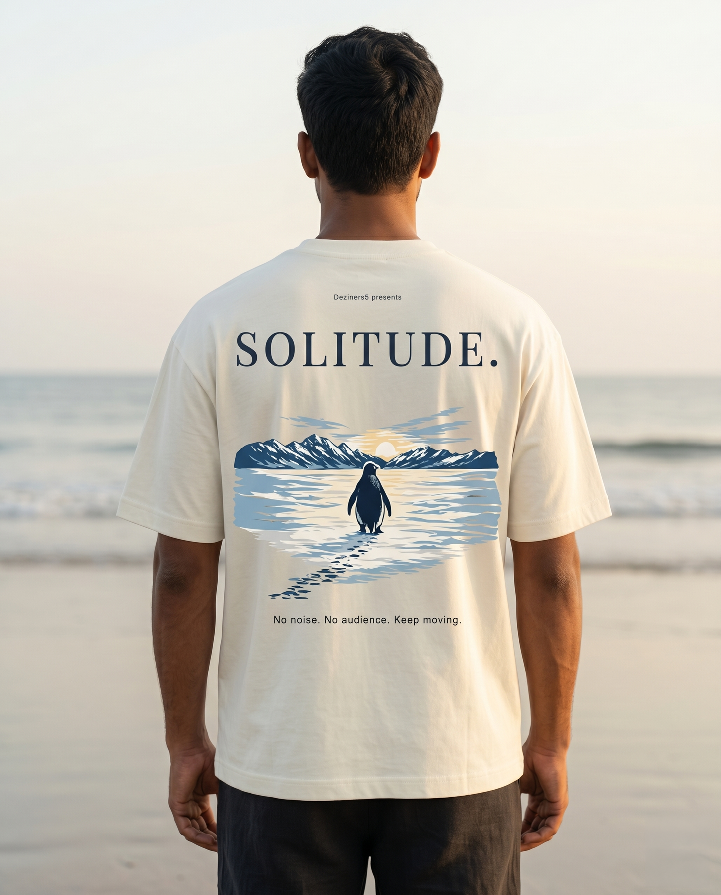 South Indian man wearing the SOLITUDE - unisex oversized graphic t-shirt in off-white on a beach at sunrise featuring inspirational penguin artwork.