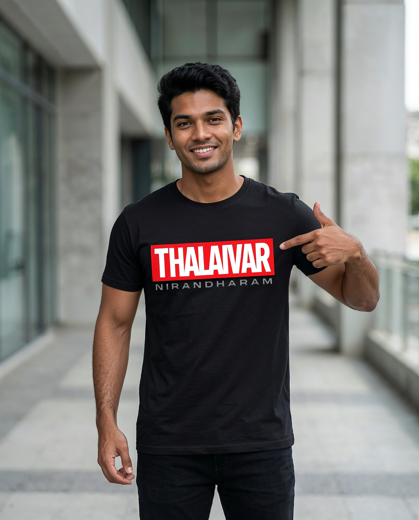 Young South Indian male wearing the THALAIVAR NIRANDHARAM men's regular-fit t-shirt in black, pointing at the front graphic print in a modern urban outdoor setting.