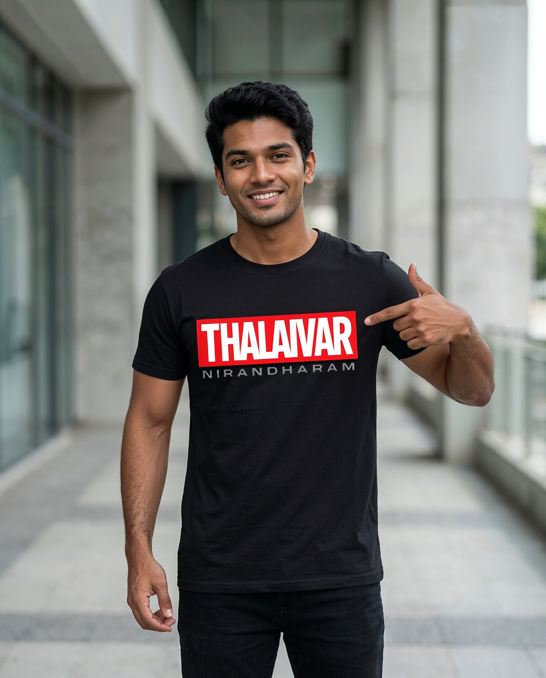 Young South Indian male wearing the THALAIVAR NIRANDHARAM men's regular-fit t-shirt in black, pointing at the front graphic print in a modern urban outdoor setting.